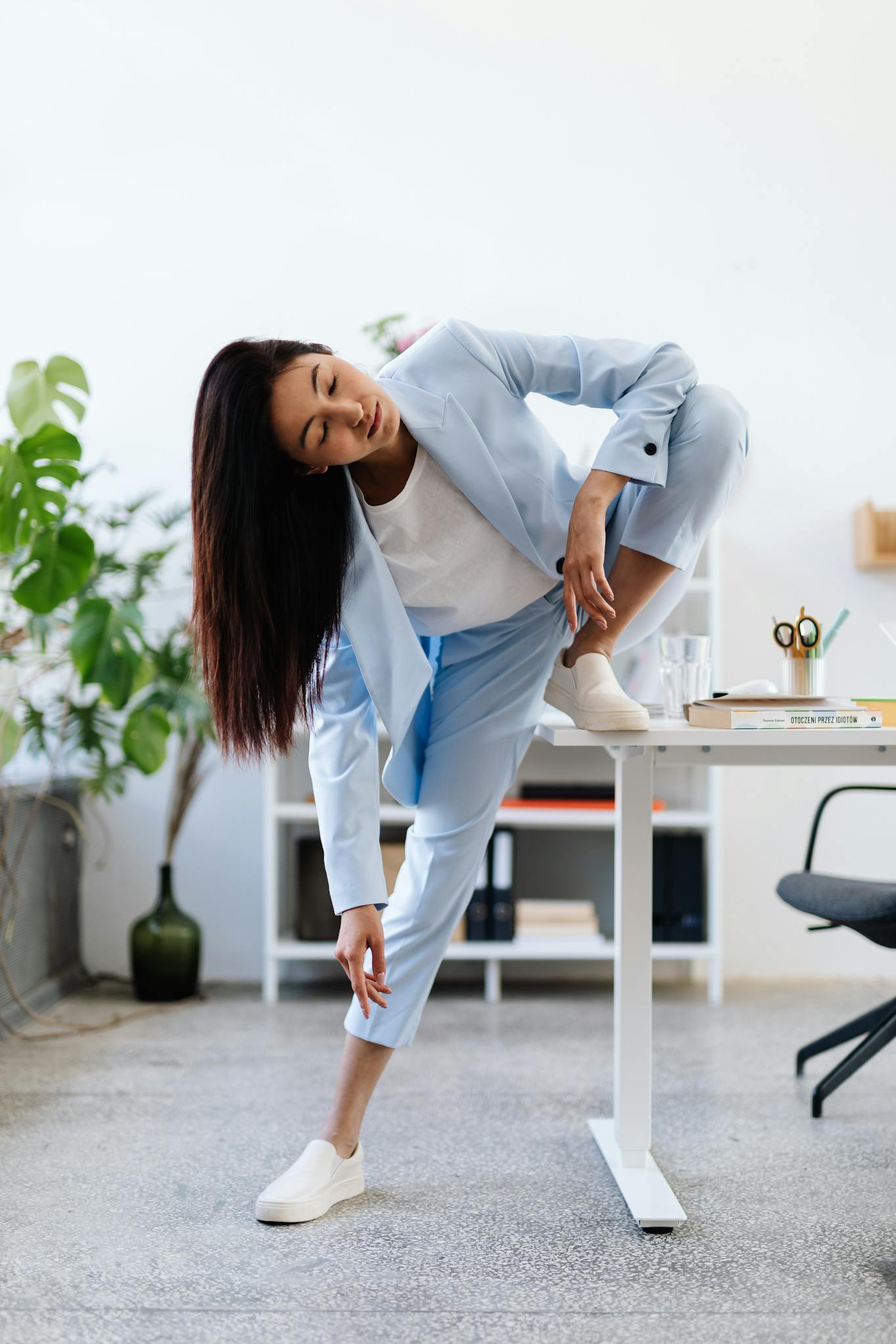 A woman in a blue suit practicing yoga poses on a desk in a bright office.