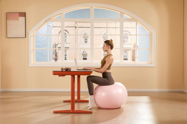 A young woman works on a laptop while sitting on an exercise ball in a bright, modern room.
