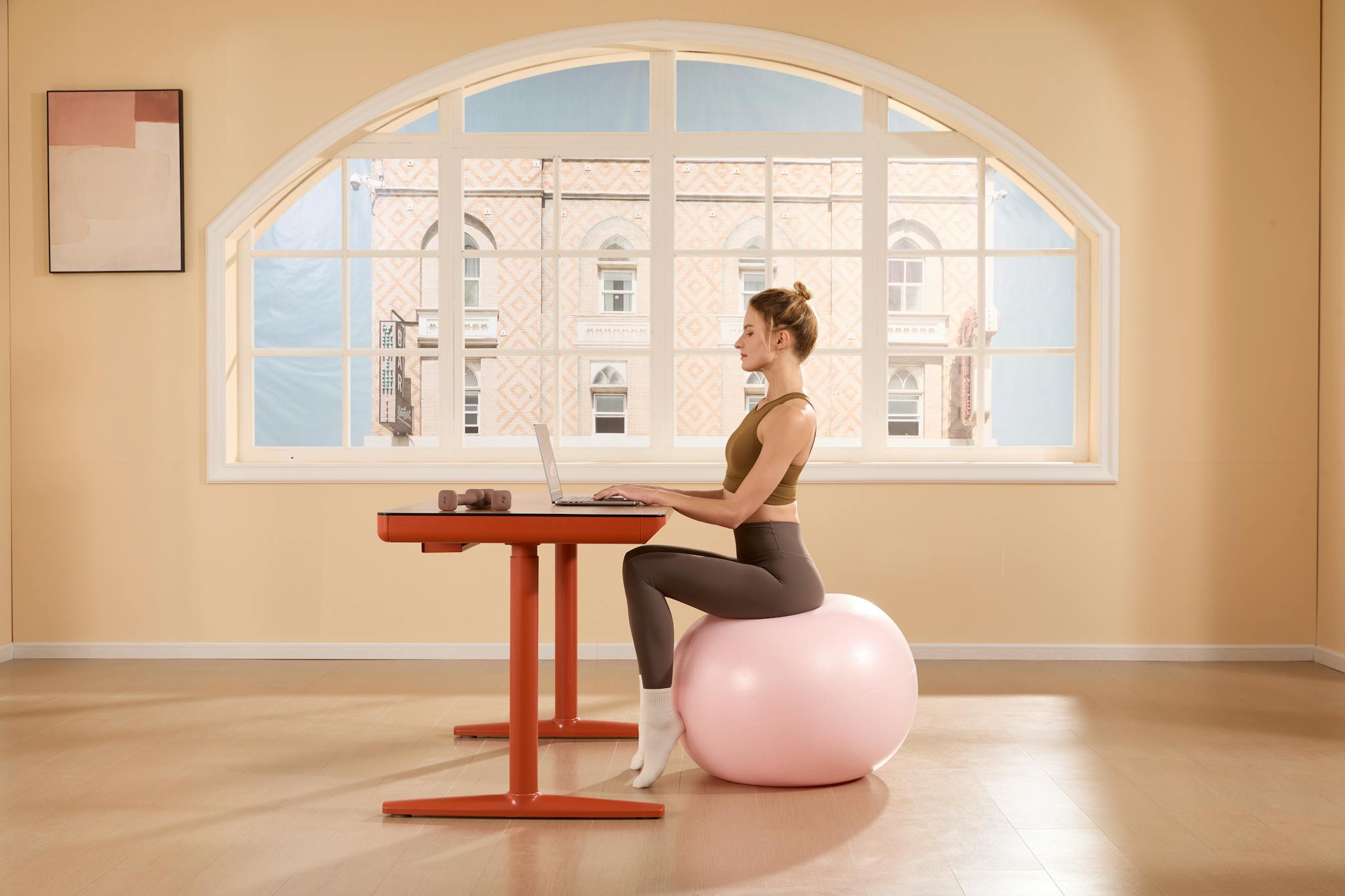 A young woman works on a laptop while sitting on an exercise ball in a bright, modern room.