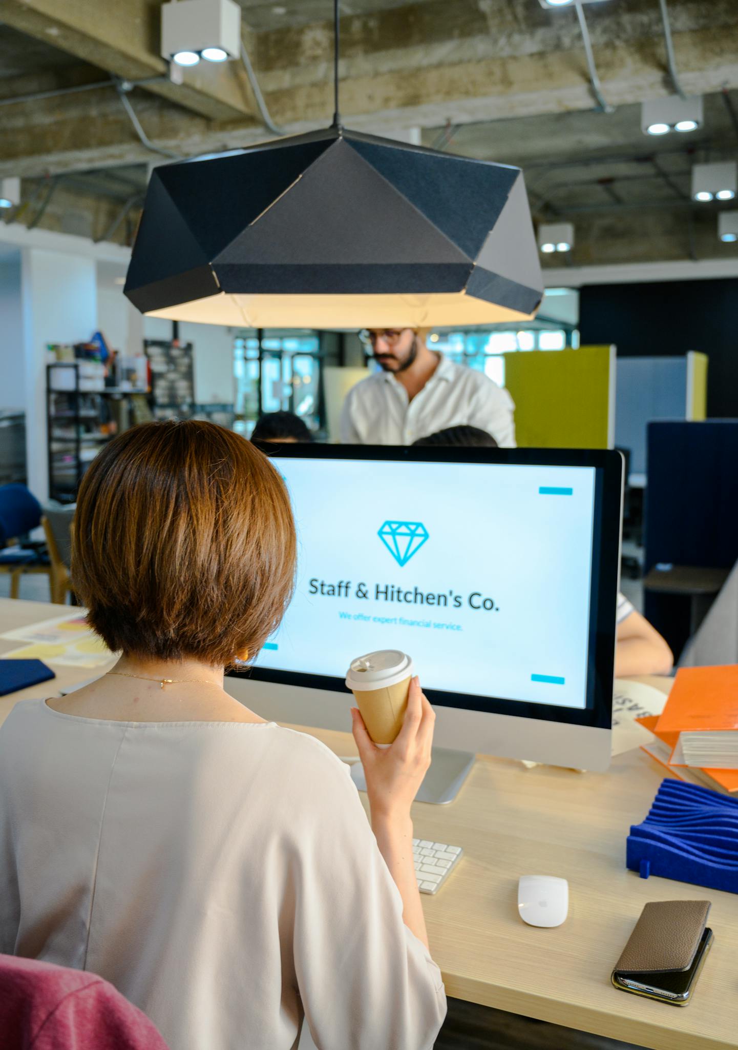 Person holding a coffee cup, sitting at a desk with a computer screen displaying 'Staff & Hitchen's Co.' in a modern office.