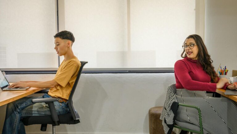 Two colleagues work opposite each other in a bright, modern office setting, engaged with laptops.